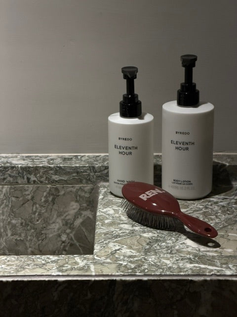 Two white pump bottles labeled ELEVENTH HOUR and REN brand with a dark red,  burgundy Bordeaux mini hairbrush resting in front.